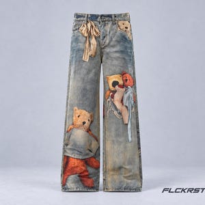 Peut inclure: Jean en denim bleu clair délavé à jambes larges avec plusieurs appliques d'ours en peluche. L'avant présente un ruban beige à la taille, une petite tête d'ours sur la poche et des ours plus grands sur les jambes. Le texte indique FLCKRSTUDIO.