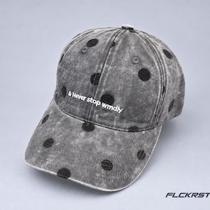 May include: A charcoal grey acid-wash baseball cap featuring a black polka dot pattern and the white embroidered text & Never stop wmdly on the front panel. The cap includes a curved brim, stitched eyelets, and a structured six-panel design.