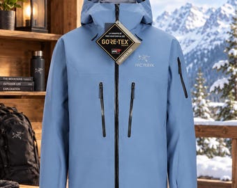May include: A light blue Arc'teryx hooded jacket with a full-length black front zipper, two vertical chest pockets with black zippers, and a small sleeve pocket. A diamond-shaped Gore-Tex tag hangs from the collar, and the brand logo is on the chest.