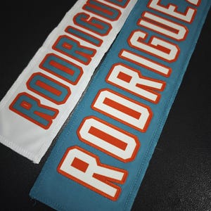 May include: Two rectangular fabric patches featuring the name RODRIGUEZ in block lettering. One patch is white with blue and orange text, while the other is light blue with white and orange text, both showing stitched borders.