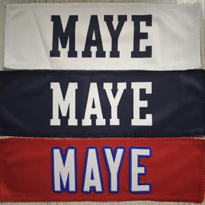 May include: Three rectangular fabric patches featuring the text MAYE in block lettering. The top patch is white with dark blue text, the middle is dark blue with white text, and the bottom is red with white text and blue outlines.