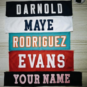 May include: Five rectangular fabric name patches with block lettering. The top patch is black with white text DARNOLD, followed by white with navy text MAYE, teal with orange text RODRIGUEZ, red with white text EVANS, and black with pink text YOUR NAME.
