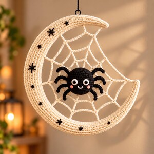 May include: A cream-colored crocheted crescent moon wall hanging featuring a central spider web pattern. A small black crocheted spider with white eyes and pink cheeks is attached to the web. The moon is decorated with small black embroidered stars.