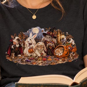 May include: A dark gray t-shirt featuring a graphic illustration of several rabbits dressed in Halloween costumes, including a vampire, a ghost, a mummy, and a witch, surrounded by pumpkins, candy, and mice on a cobblestone path at night.