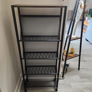 May include: A tall, rectangular storage rack featuring a dark brown wooden top shelf and a black metal frame. The unit includes four slanted, black mesh shelves designed for organization, supported by a sturdy, vertical black metal structure.