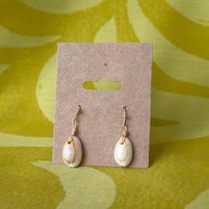May include: A pair of drop earrings featuring small, white, oval-shaped cowrie shells with natural orange-yellow markings. Each shell is securely attached to a gold-toned metal hook by a thin, coiled gold-toned wire wrap, displayed on a brown card.