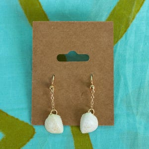 May include: A pair of dangle earrings featuring small, off-white seashell charms suspended from thin gold-toned metal chains and hook-style ear wires, displayed on a rectangular brown cardboard jewelry backing card.