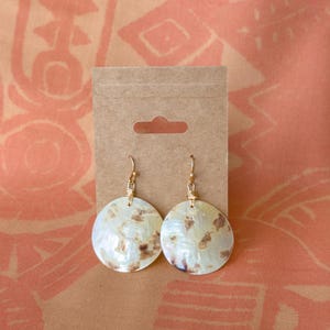 May include: A pair of round, flat, iridescent shell earrings with brown mottled patterns. Each earring features a gold-toned wire hook and a small wire-wrapped loop at the top, displayed on a rectangular brown kraft paper card with a cutout tab.
