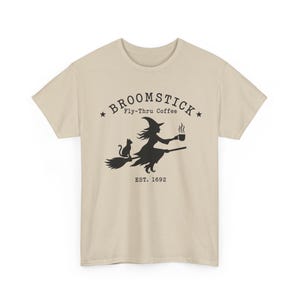 May include: A beige short-sleeved t-shirt featuring a black graphic of a witch riding a broomstick with a cat and a steaming coffee mug. The text reads BROOMSTICK Fly-Thru Coffee EST. 1692, framed by two small stars above the central illustration.