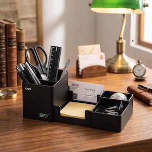 May include: A black plastic desk organizer featuring a tall compartment for pens, scissors, and a ruler, a central slot for business cards and sticky notes, and a smaller side bin for paper clips. The front displays the white text Deli.