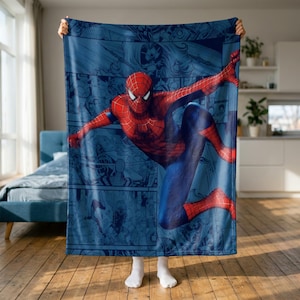 May include: A rectangular throw blanket featuring a large, central graphic of the superhero Spider-Man in a dynamic leaping pose. The background consists of a blue-toned comic book panel collage. The character wears a red and blue suit with a web pattern.