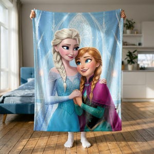 Puede incluir: Una manta rectangular de forro polar con una ilustración impresa de dos personajes animados con cabello rubio y castaño rojizo. El personaje rubio viste un vestido azul claro, mientras que el personaje de cabello castaño viste un vestido verde.