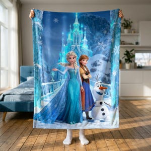 Puede incluir: Una manta rectangular de forro polar con un estampado colorido de Elsa, Anna y Olaf de Frozen. El diseño muestra un castillo de hielo azul brillante en un paisaje nevado con copos de nieve. Los personajes están situados en primer plano.
