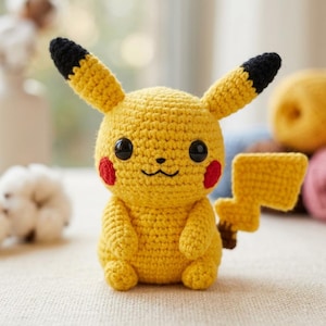 May include: A yellow crocheted stuffed toy featuring long ears with black tips, round black eyes, small red circular cheeks, and a lightning bolt-shaped tail. The character has a small embroidered mouth and sits in an upright position.