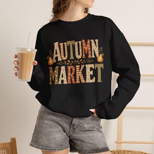 May include: A black long-sleeve sweatshirt featuring the text AUTUMN MARKET in a decorative font with various patterns including plaid, floral, and solid colours. The design includes two pears and leaf accents in shades of orange, brown, and cream.