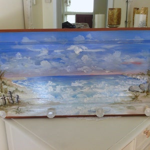 May include: A wooden framed painting of a seascape with a blue sky, white clouds, and a beach with waves crashing on the shore. The painting is mounted on a wall with clear plastic knobs.