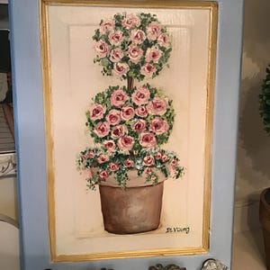 May include: A framed painting of a topiary with pink roses in a brown pot. The painting is set in a light blue frame with gold trim. The painting is signed "DL Young" in the bottom right corner. The frame has a decorative handle and crystal knobs.