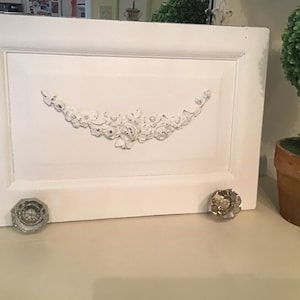 May include: White cabinet door with a decorative floral design and two crystal knobs. The door has a raised panel and a detailed, curved floral embellishment. The knobs are clear and faceted.