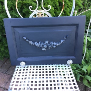 May include: A dark blue painted wooden door with a decorative floral design and two silver door knobs. The door is hanging on a white metal chair.
