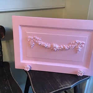 May include: A light pink, rectangular wooden panel with a raised floral and ribbon design. The panel has a decorative border and is supported by two small, rose-shaped feet. The panel is resting on a dark wooden chair.