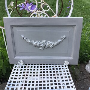 May include: A gray painted wooden cabinet door with a silver floral design and two silver knobs.