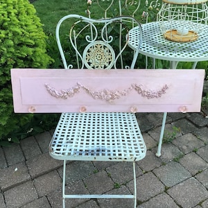 May include: A pink wooden wall-mounted shelf with three decorative knobs. The shelf has a raised floral design with a scalloped edge.