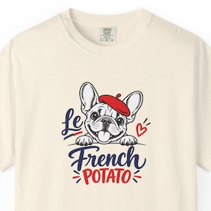 Könnte beinhalten: Ein cremefarbenes Kurzarm-T-Shirt mit einer Grafik einer französischen Bulldogge, die ein rotes Barett trägt. Der Text lautet Le French POTATO in blauer und roter Schrift, ergänzt durch ein kleines rotes Herzsymbol.