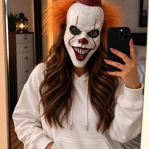 Pode incluir: Uma máscara de palhaço branca com superfície texturizada, nariz vermelho, linhas vermelhas pintadas saindo dos olhos e um sorriso largo com dentes afiados. A máscara tem cabelo sintético laranja brilhante nas laterais e uma parte superior moldada em vermelho escuro.
