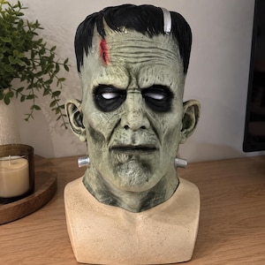 Puede incluir: Un busto del monstruo de Frankenstein con piel verde pálido, cabello negro con un acento plateado y una cicatriz roja en la frente. La figura tiene ojos hundidos, arrugas faciales prominentes y pernos de color plateado en el cuello.