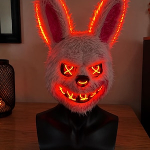 Puede incluir: Una máscara de conejo de peluche con orejas largas y un exterior de piel sintética texturizada en color blanco roto. La máscara está iluminada por tiras de luz de neón rojo brillante que delinean las orejas, los ojos en X, la nariz y una sonrisa.