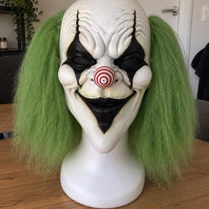 Pode incluir: Uma máscara de palhaço de cabeça inteira com um rosto branco pálido, marcas pretas profundas nos olhos, um sorriso preto largo e um nariz proeminente com um padrão de alvo vermelho e branco. Possui cabelo verde texturizado em ambos os lados.