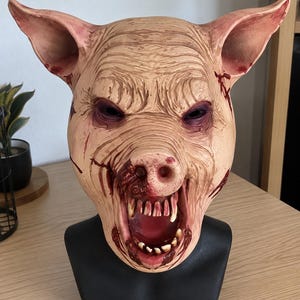 Könnte beinhalten: Eine realistische Schweinemaske mit strukturierter Haut in Beige- und Rosatönen, großen spitzen Ohren, tief liegenden dunklen Augen und einem offenen Maul, das spitze, verfärbte Zähne zeigt. Die Maske ist mit roten blutartigen Spritzern versehen.