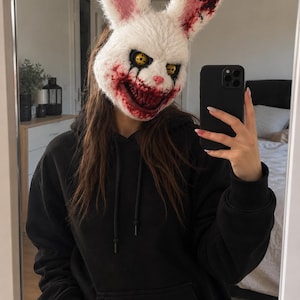 Puede incluir: Una máscara de conejo blanca y peluda con orejas largas, ojos amarillos con pupilas negras y una amplia sonrisa dentada con salpicaduras de color rojo sangre alrededor de la boca y la oreja. Se lleva con una sudadera negra y un smartphone.