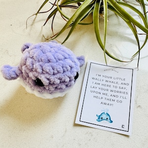 May include: A purple crocheted whale plush toy with a white belly and black eyes. The toy is sitting on a white surface next to a card that reads "WALLY WHALE I'M YOUR LITTLE WALLY WHALE, AND I AM HERE TO SAY, LAY YOUR WORRIES UPON ME, AND I'LL HELP THEM GO AWAY!"