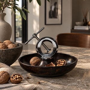 May include: A dark brown circular bowl featuring a central chrome-plated metal nutcracker mechanism. The bowl contains several whole and cracked walnuts. The nutcracker has a T-shaped handle and a threaded screw design for cracking shells.