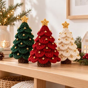 May include: Three crocheted Christmas tree decorations in dark green, red, and cream. Each tree features layered, scalloped branches, a brown trunk, and a small yellow crocheted star topper. Small, shiny gold beads are attached to the branches.