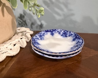 May include: A small, round ceramic saucer featuring a white center and a scalloped edge decorated with a blue floral pattern and a thin gold-toned rim. The saucer has a slightly raised, wavy border and a smooth, glossy finish.