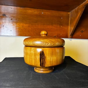 Puede incluir: Un cuenco redondo de madera de color marrón dorado con tapa y acabado pulido. El cuenco presenta una prominente inclusión vertical oscura de veta natural en el lateral y se asienta sobre una pequeña base. Una tapa de madera a juego con pomo.