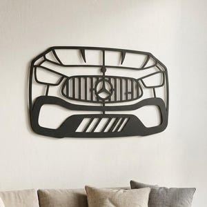 Op de afbeelding: Een matzwart metalen wanddecoratie in de vorm van de voorgrille en bumper van een Mercedes-Benz voertuig. Het ontwerp toont het iconische driepuntige ster-embleem gecentreerd tussen verticale lamellen, geflankeerd door gestileerde koplampen.