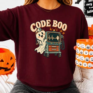 Puede incluir: Una sudadera granate de cuello redondo con un gráfico de un fantasma con estetoscopio y espejo frontal, junto a un monitor médico que muestra un latido. El texto CODE BOO aparece impreso en una fuente curva de color crema sobre el gráfico.