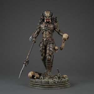May include: A detailed Predator figurine standing on a rocky base with skulls. The figure wears brown armor, has long dark hair, and holds a long silver spear in one hand and a skull with a spinal column in the other. The base features scattered skulls.