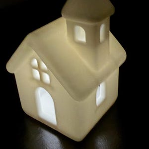 May include: A small, off-white ceramic house-shaped luminary with a steeple, featuring cut-out windows and a door that emit a soft, warm internal glow. The structure has a gabled roof and a square base, illuminated from within against a dark background.
