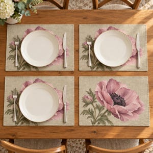 May include: A set of four rectangular placemats featuring a light beige background with a large, detailed illustration of a pink poppy flower and green leaves. Three placemats are set with a white ceramic plate, a silver fork, and a silver knife.