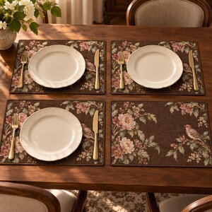 May include: Four rectangular placemats with a dark brown background featuring a floral and bird pattern in shades of pink, green, and cream. Three placemats are set with a white ceramic plate, a gold-toned fork, and a gold-toned knife.