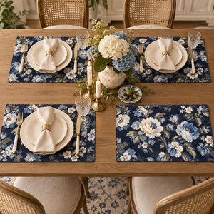 Peut inclure: Un ensemble de sets de table rectangulaires avec un fond bleu foncé et un motif floral blanc et bleu clair. La table est dressée avec des assiettes crème, des serviettes en lin beige avec des ronds de serviette dorés, des couverts dorés et des verres.