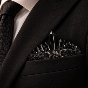 May include: A black pocket square accessory featuring a cluster of dark, pointed crystal-like structures. The piece is adorned with a central metallic pentagram symbol flanked by two crescent moon shapes, all set against a textured black base.