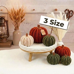 May include: A collection of six knitted pumpkins in cream, rust orange, and olive green, each topped with a cinnamon stick stem. The pumpkins are displayed on a white wooden pedestal stand, with a sign clearly displaying the text 3 Size.