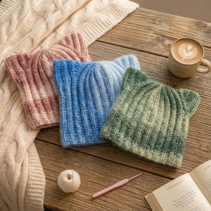 Pode incluir: Três gorros de malha canelada com silhuetas de orelhas de gato, feitos com fio matizado em tons de rosa antigo, azul celeste e verde floresta. Cada gorro apresenta um padrão de textura vertical e uma aparência macia e felpuda.