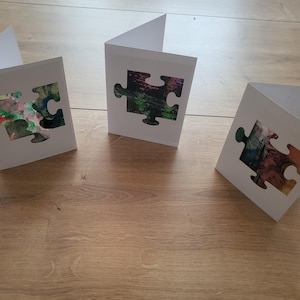 Könnte beinhalten: Drei weiße, gefaltete Grußkarten, jede mit einer mittigen Stanzung in Form eines Puzzleteils. Die Ausschnitte geben den Blick auf abstrakte, farbenfrohe gemalte Muster in Grün-, Blau-, Rosa- und Orangetönen frei, die durch die Karten sichtbar sind.
