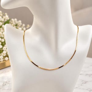 May include: A gold-toned herringbone chain necklace featuring a flat, woven link design that rests in a smooth curve. The necklace has a polished, reflective surface that catches the light, creating a consistent metallic sheen across the entire length.
